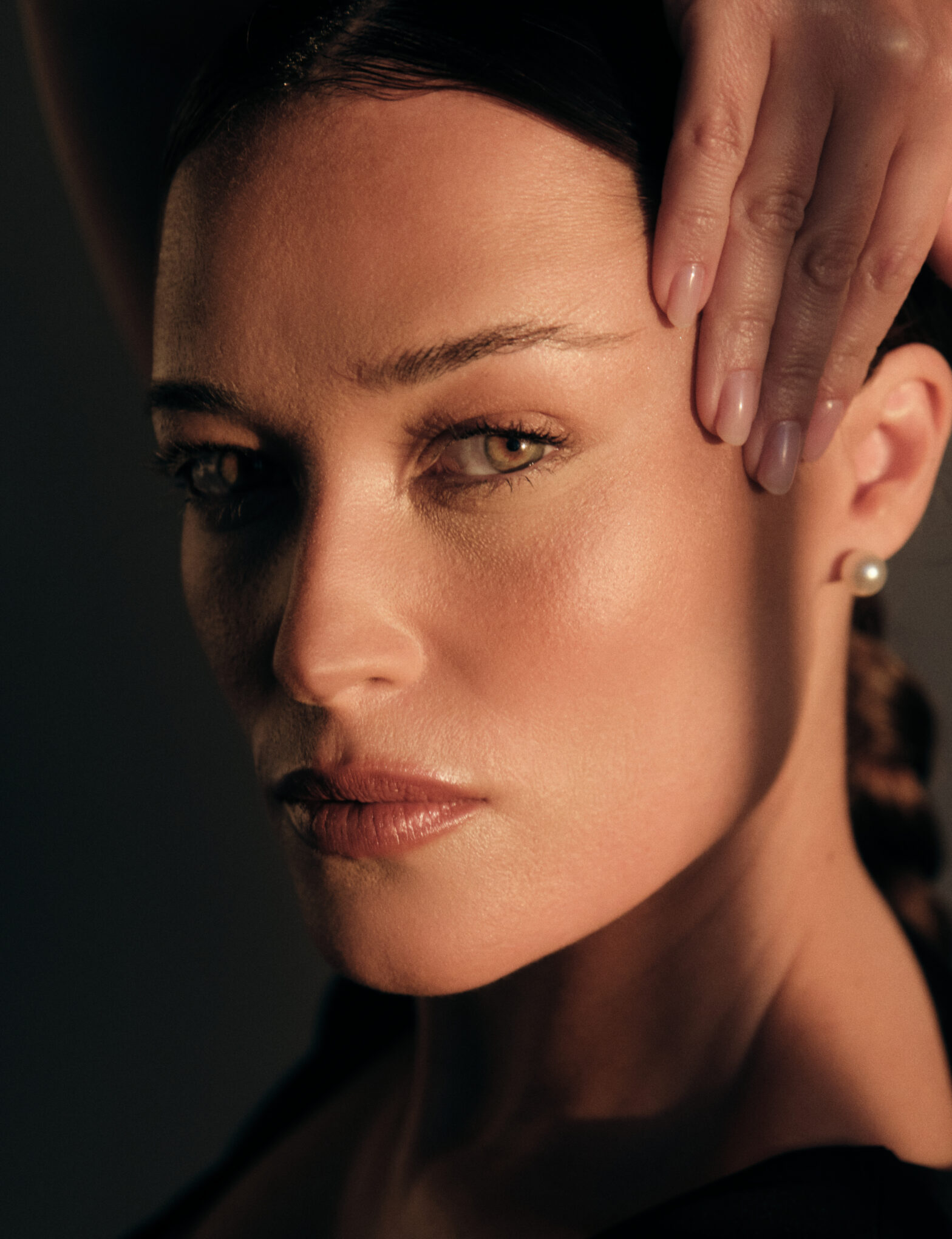 Close-up portrait of a woman with luminous skin touching her temple in warm natural light.