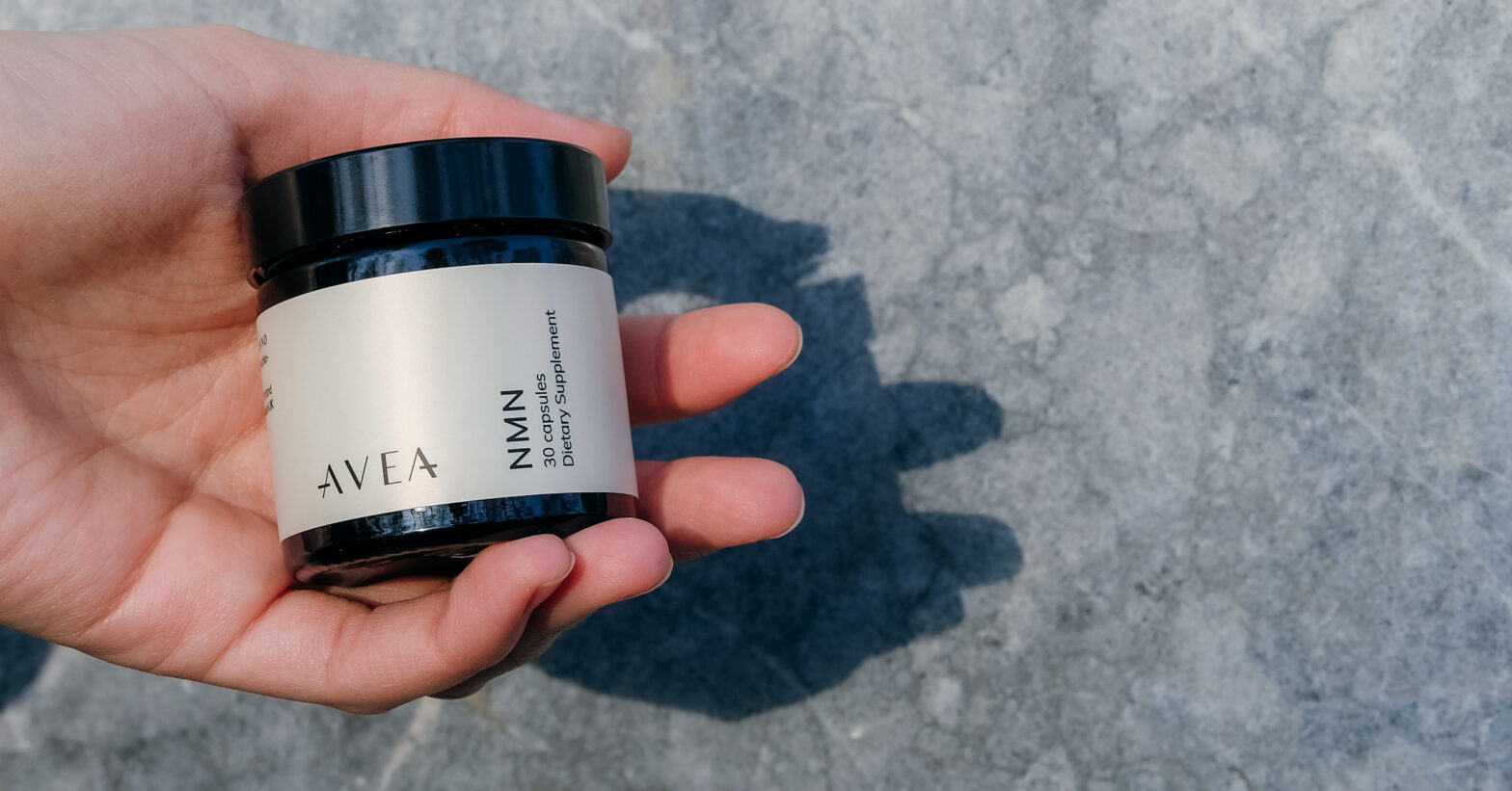 A hand holding a small AVEA NMN supplement jar with a white label and black lid against a gray stone background in natural sunlight.
