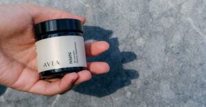 A hand holding a small AVEA NMN supplement jar with a white label and black lid against a gray stone background in natural sunlight.
